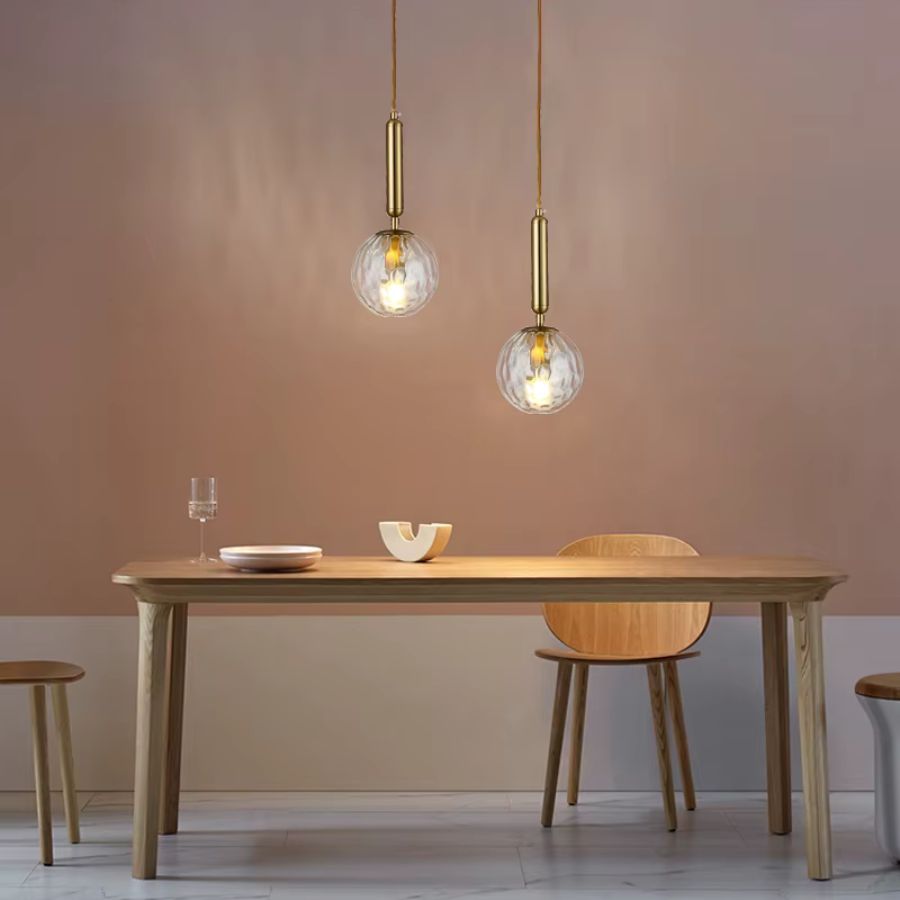 Deux suspensions en verre transparent à tiges dorées au-dessus d’une table, dans une salle à manger sobre et chaleureuse