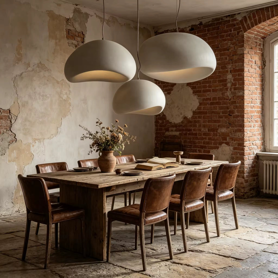 Trois suspensions blanches à dômes asymétriques illuminent une table en bois brut dans une salle à manger rustique aux murs de briques.