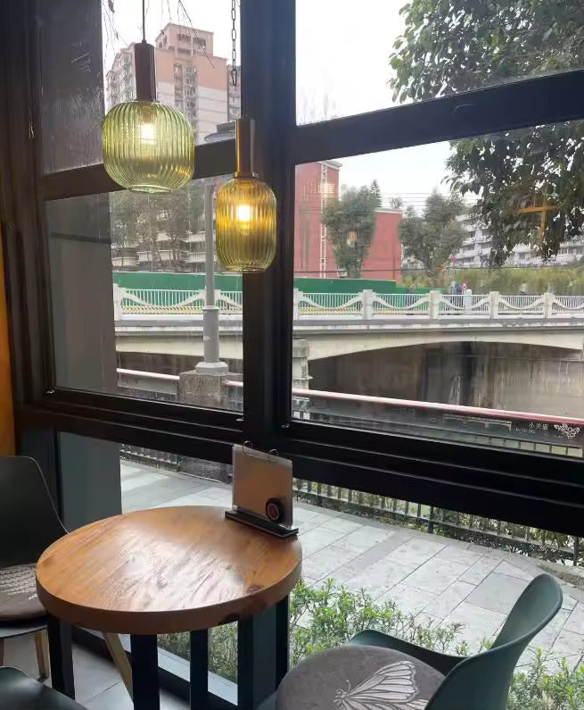 Deux suspensions en verre cannelé vert avec finitions dorées suspendues près d’une grande fenêtre dans un café. Ces luminaires élégants éclairent une table ronde et créent une ambiance chaleureuse avec vue sur l’extérieur.