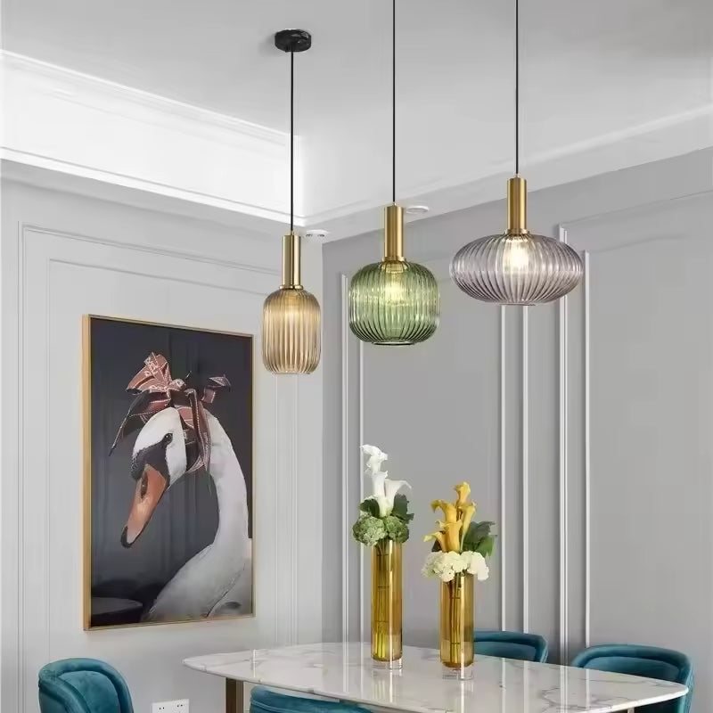 Trois suspensions en verre cannelé aux teintes ambrée, verte et fumée avec finitions dorées, installées dans une salle à manger moderne. Ces luminaires décoratifs créent une ambiance élégante et chaleureuse au-dessus de la table.