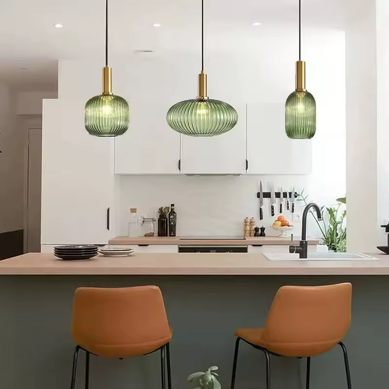 Trois suspensions en verre cannelé vert avec finitions dorées installées au-dessus d’un îlot de cuisine moderne. Ces luminaires élégants diffusent une lumière douce et apportent une touche raffinée à l’espace.